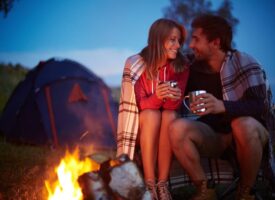 Young couple enjoying warm drinks covered in a blanket by a campfire at dusk.